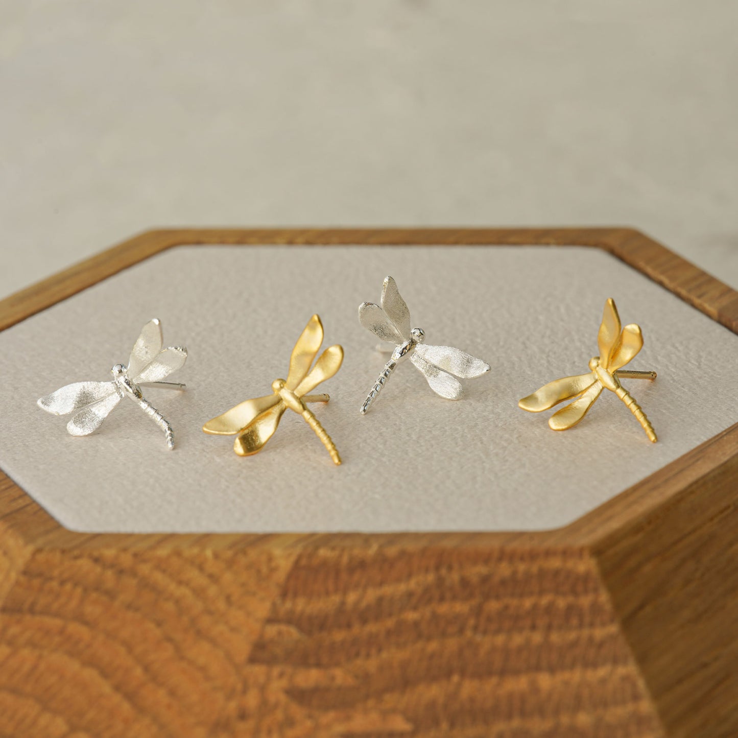Lineup of gold and silver dragonfly stud earrings displayed on a hexagonal wood tray, showing color options and natural design