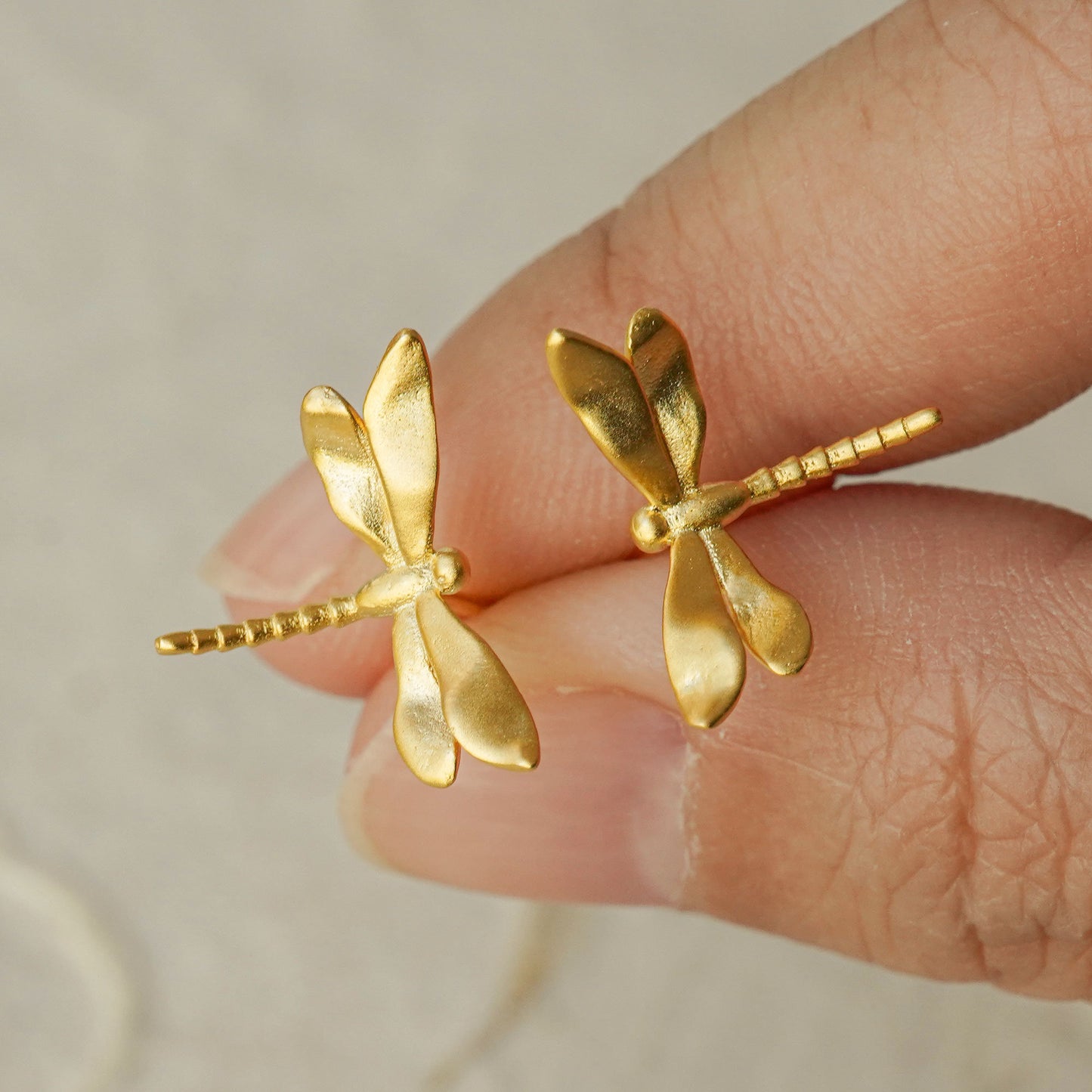 Close-up of two gold dragonfly stud earrings held between fingers, showing their elegant wings and shiny finish