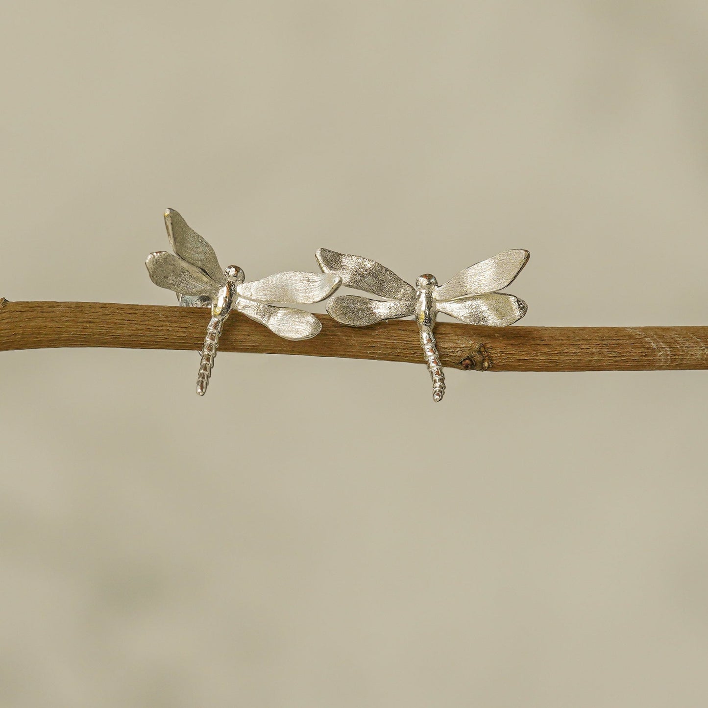 Silver dragonfly stud earrings displayed on a wooden branch, mimicking a natural resting pose with elegant detail