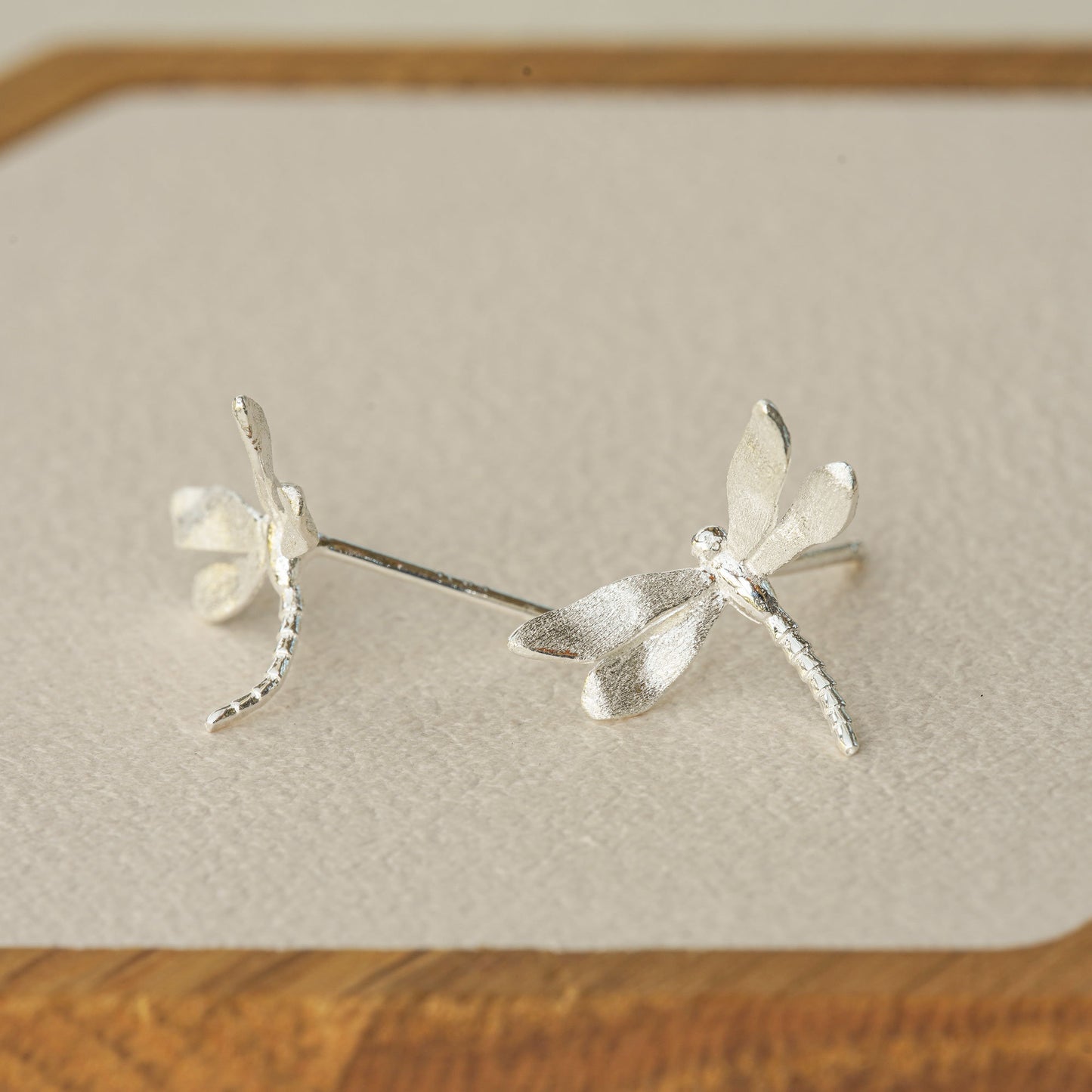 Silver dragonfly stud earrings displayed on a wooden tray, highlighting their delicate shape and textured finish