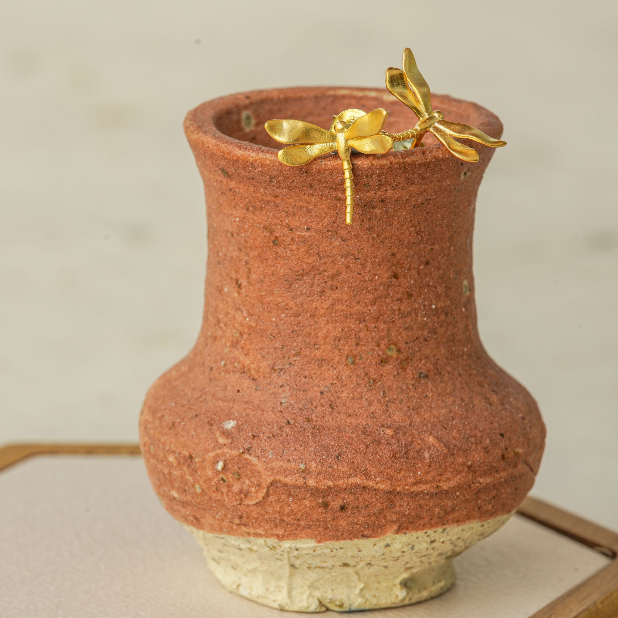 Gold dragonfly stud earrings resting on the rim of a rustic clay vase