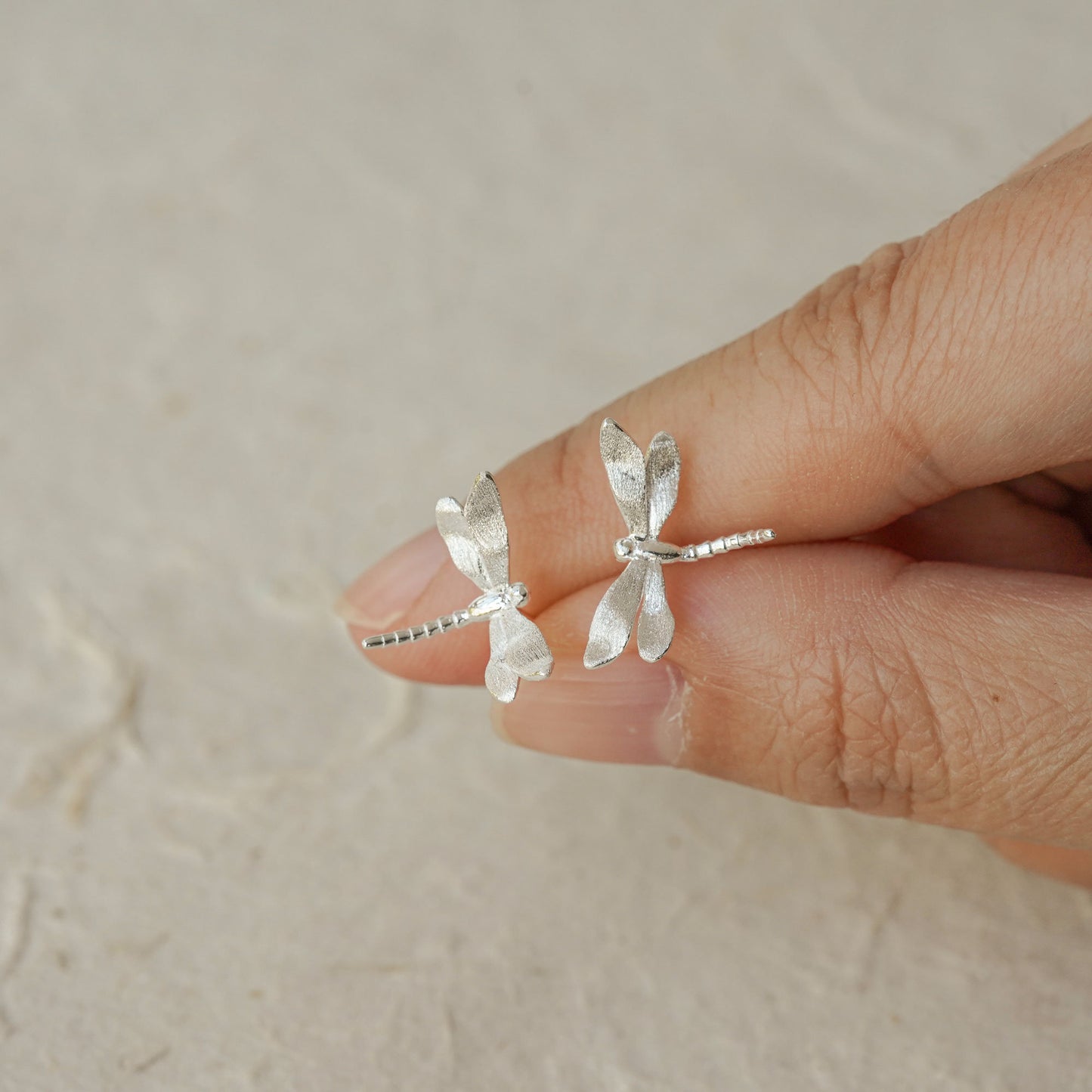 Close-up of two silver dragonfly stud earrings held between fingers, highlighting their textured wings and lightweight design