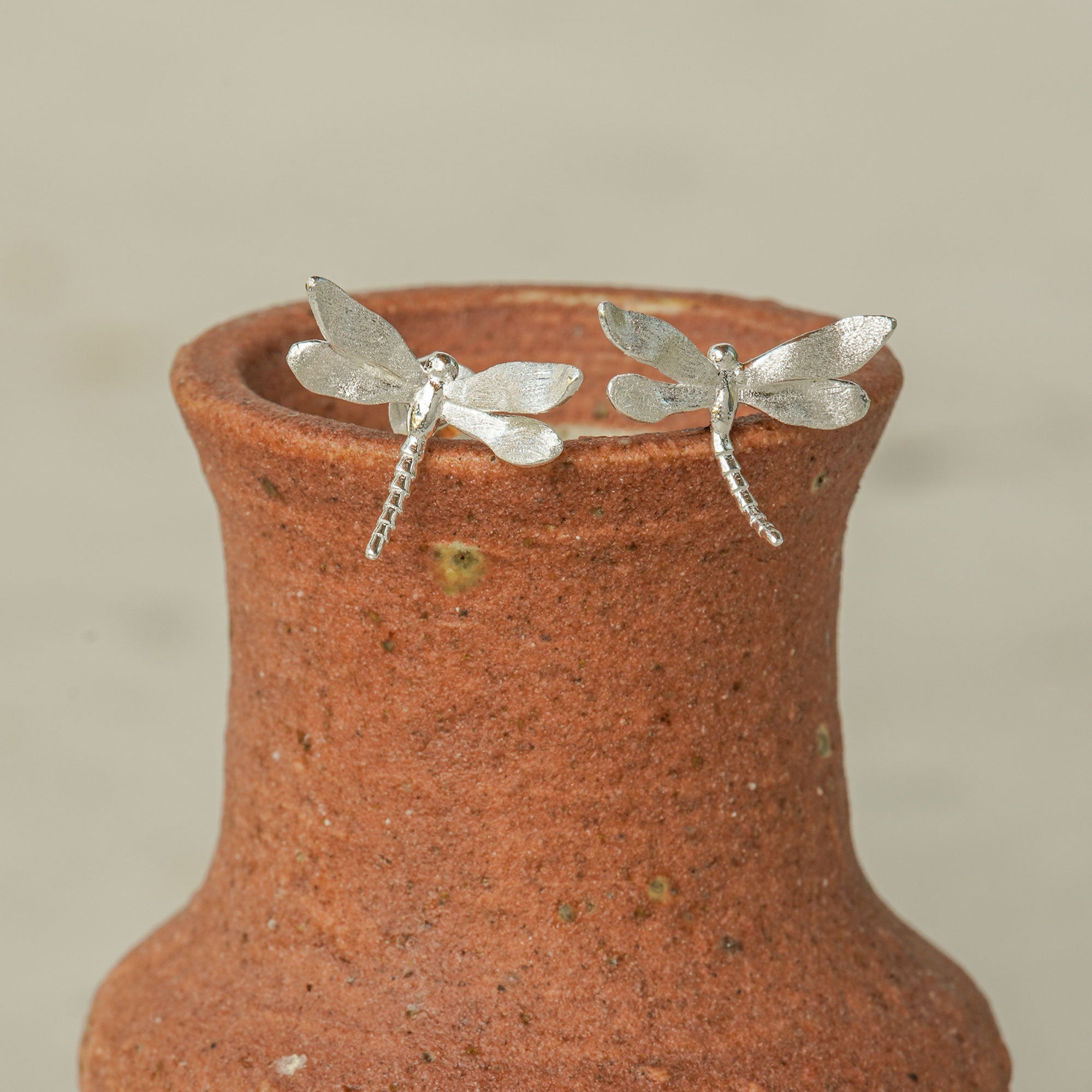 Silver dragonfly stud earrings displayed on the rim of a rustic clay vase