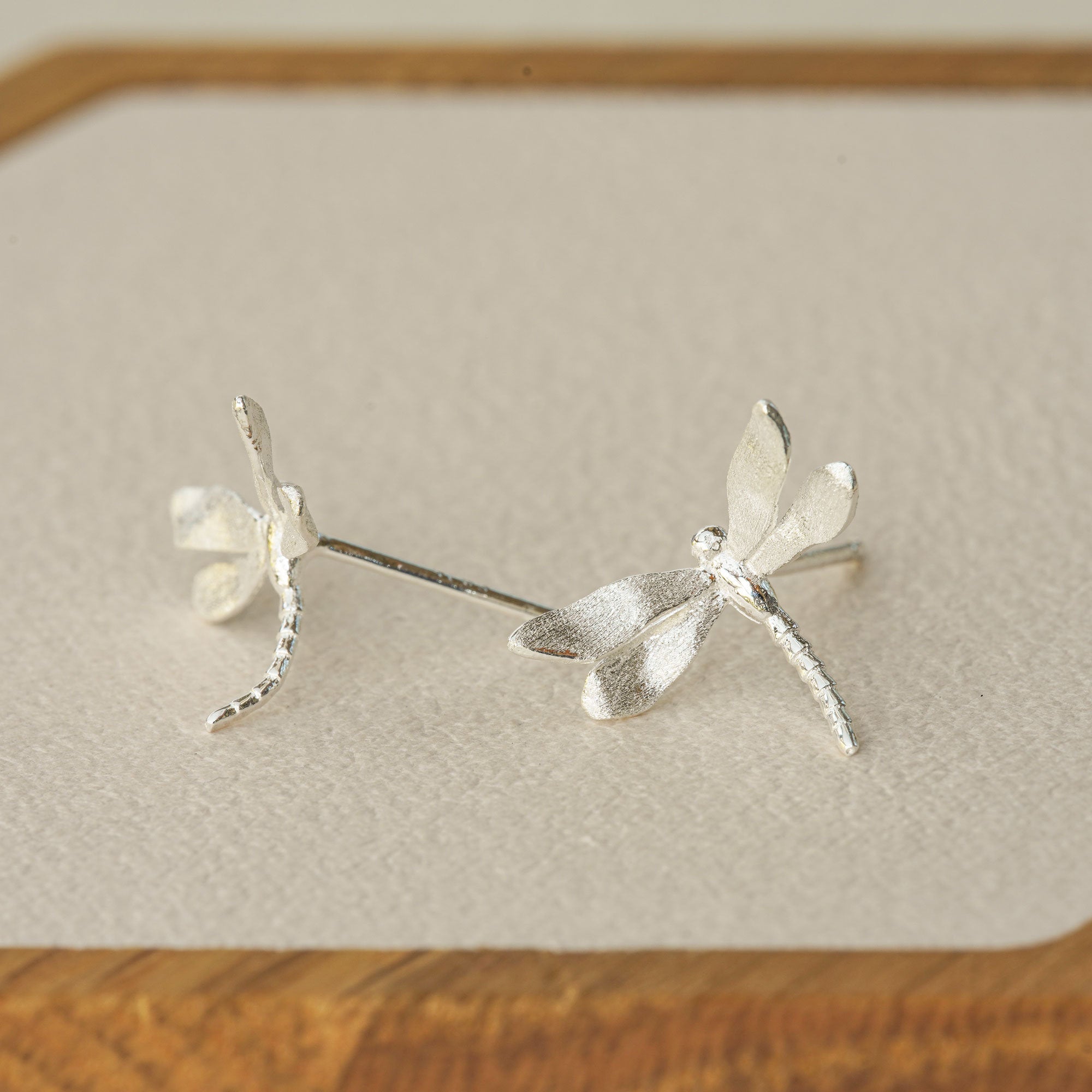 Silver dragonfly stud earrings displayed on a wooden tray, highlighting their delicate shape and textured finish